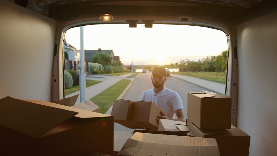 A white moving van with the branding 'MOVING COMPANY LOCAL & LONG DISTANCE' on its side parked on a city street in front of a multi-story brick building with large arched windows. The van's driver, we