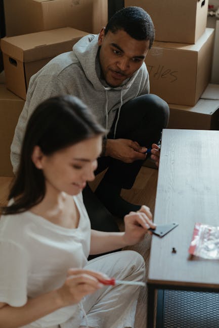 A young man and woman engaged in house moving preparations inside a room with wooden flooring, surrounded by stacked cardboard boxes, some labeled and sealed with packing tape. The woman, seated at a