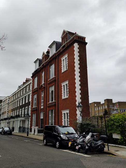 A multi-story red brick building with white detailing on the corners and window frames, situated on a street with parked vehicles including a black van and several scooters, under an overcast sky. The