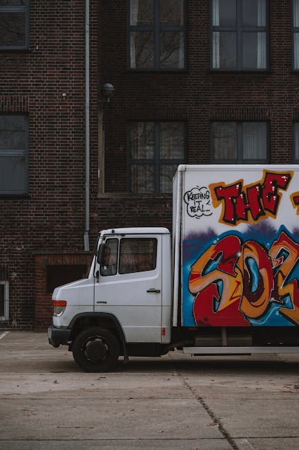 A white U-Haul moving truck parked on a gravel surface in an open outdoor area with a coastal landscape in the background. The truck's cargo area features the U-Haul logo and slogan 'MAKES MOVING EASI